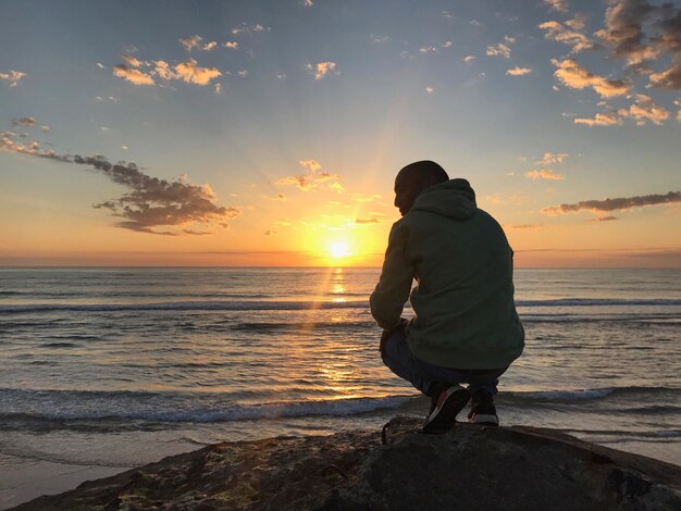 Foto vista posteriore a tutta lunghezza di un uomo accovacciato su una roccia sulla spiaggia durante il tramonto