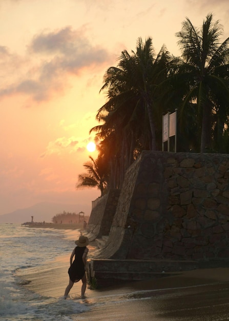 Задний вид на полную длину женщины, идущей по пляжу напротив неба во время захода солнца