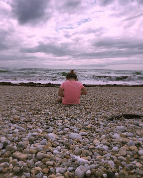 Rear view of girl sitting at beach against cloudy sky during sunset