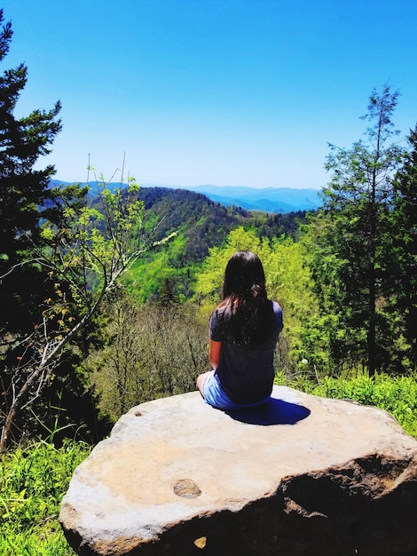 Foto vista posteriore di una ragazza seduta su una roccia contro un cielo blu chiaro durante una giornata di sole