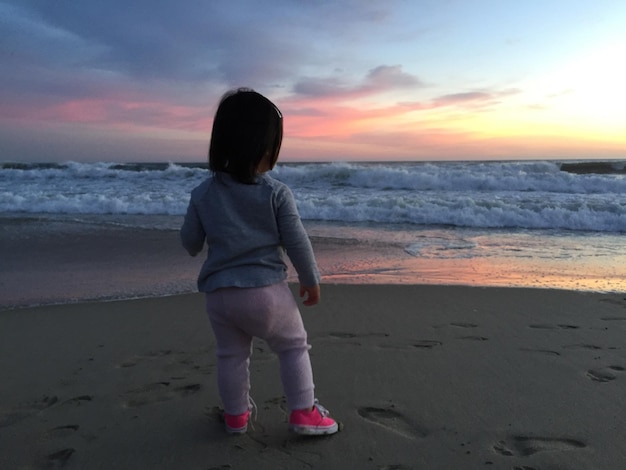 Rear view of girl standing at beach against sky during sunset