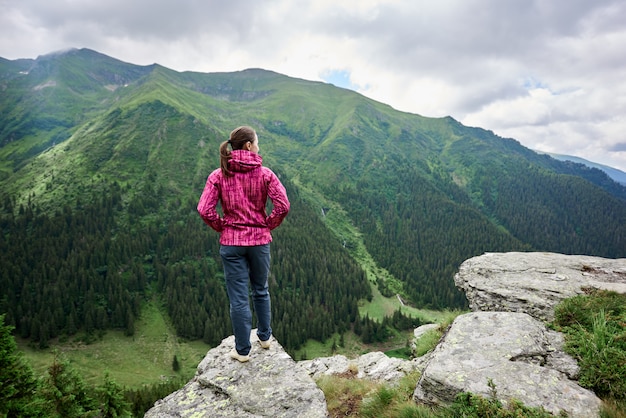 Rear view of a girl standing on a cliff in front of her opening a mesmerizing landscape of mighty green mountains and clouds above them