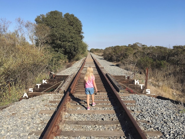 Foto vista posteriore di una ragazza che cammina sul binario ferroviario contro un cielo blu limpido