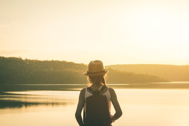 Rear view of girl with backpack standing on lakeshore during sunset