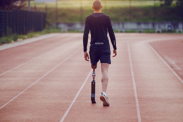 Rear view of handsome caucasian handicapped young man with artificial leg and dressed in shorts and sweatshirt walking on racetrack.