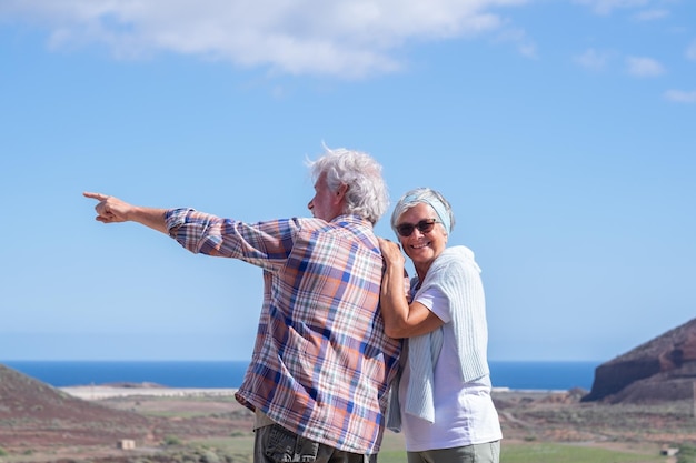 Вид сзади на счастливую активную пожилую пару, гуляющую в горах, наслаждающуюся здоровым образом жизни и солнечным днем. Два улыбающихся пожилых человека во время отдыха на горизонте над водой
