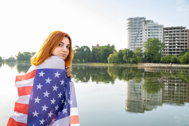 Vista posteriore di una ragazza dai capelli rossi felice con la bandiera degli stati uniti sulle spalle giovane donna positiva che celebra il giorno dell'indipendenza degli stati uniti