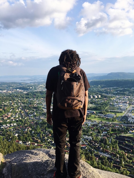rear view of hiker standing on cliff while looking at cityscape against sky