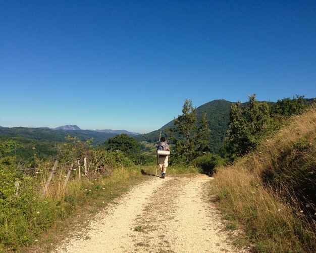 Foto vista posteriore di un escursionista che cammina su una strada di terra contro un cielo blu limpido