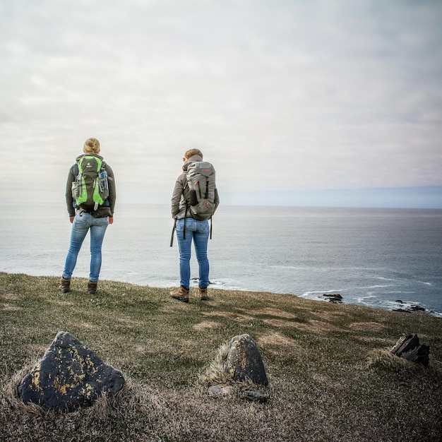 rear view of hikers standing on hill by sea against cloudy sky
