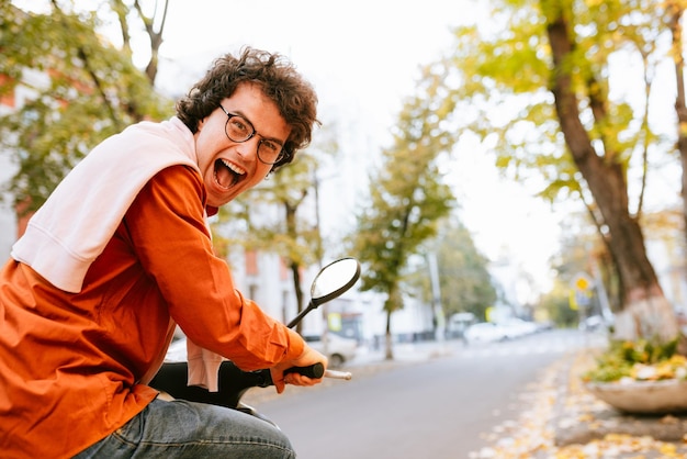 Photo rear view image of overjoyed happy male drives scooter dressed in casual outfit and transparent eyeglasses with curly hair has joyful expression reaches destination on fast transport enjoys speed