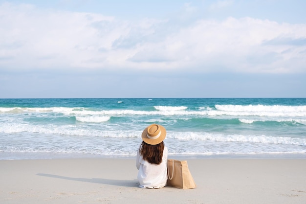 푸른 하늘을 배경으로 해변에 앉아 있는 모자와 가방을 가진 여성의 후면 이미지