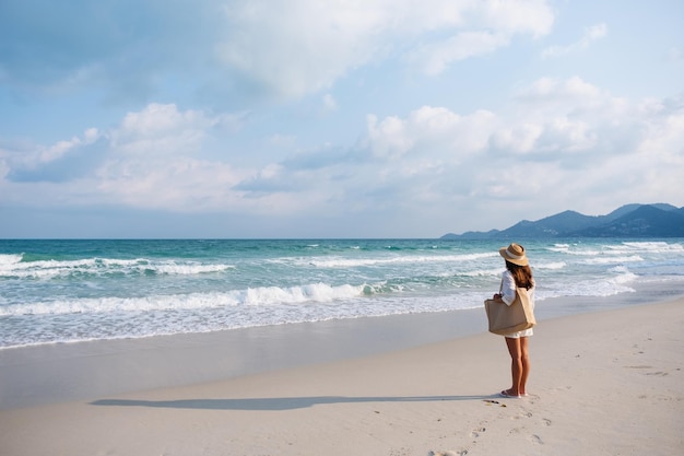 Rear view image of a woman with hat and bag strolling on the beach with blue sky background