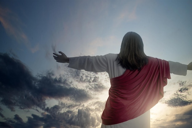 Rear view of Jesus Christ raised hands and praying to god with a dramatic sky background