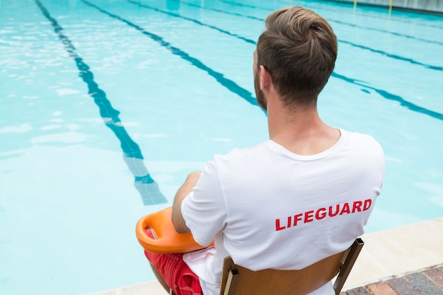 Rear view of lifeguard sitting on chair with rescue buoy at poolside