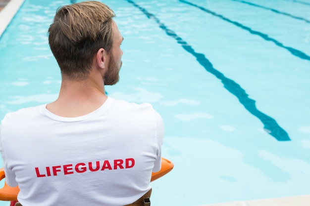 Rear view of lifeguard sitting on chair with rescue buoy at poolside