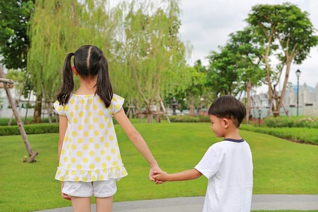 Rear view of little boy and girl child with hand in hands stand in the garden. Back view of Sister and brother clasped in the summer park.