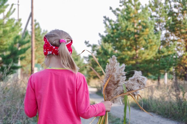 Rear view of a little girl in a pink blouse holding a bouquet of yellow stems in her hands, walking along the road. Blurred background. The idea of learning through environmental studies