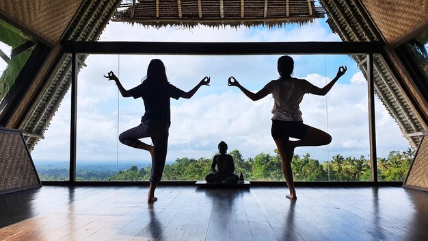 Rear view of little girl with her mother doing yoga and meditation by standing on one foot near a Buddha statue with blue sky view background at morning time