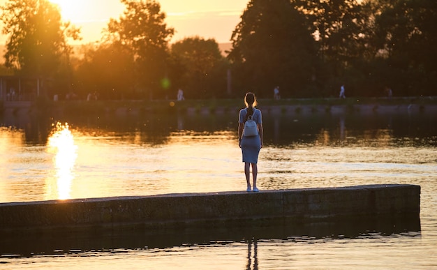 Rear view of lonely woman standing alone on lake shore on warm evening. Solitude and relaxing in nature concept.