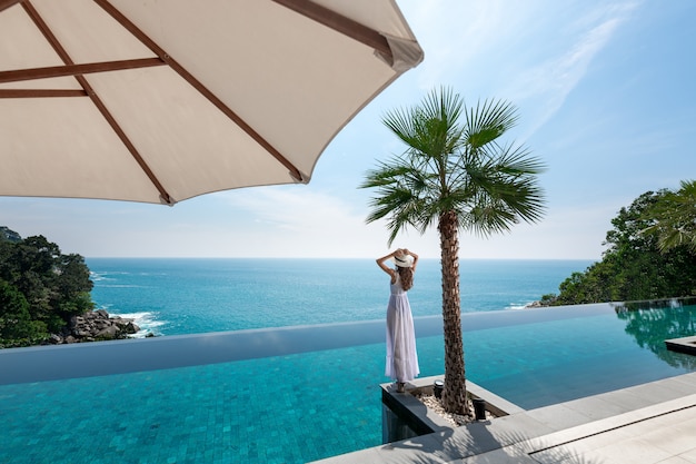 Rear view: Luxurious lady in a white dress and hat posing near a palm tree at the edge of an endless pool