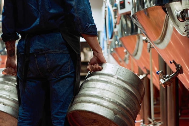 Vista posteriore del lavoratore maschio del moderno impianto di produzione di birra in abiti da lavoro che trasportano due cisterne mentre si muove lungo la fila di enormi serbatoi di acciaio