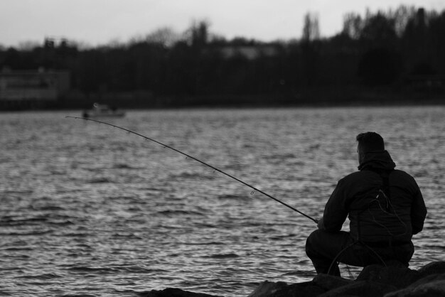 Photo rear view of man fishing while sitting on rock by lake