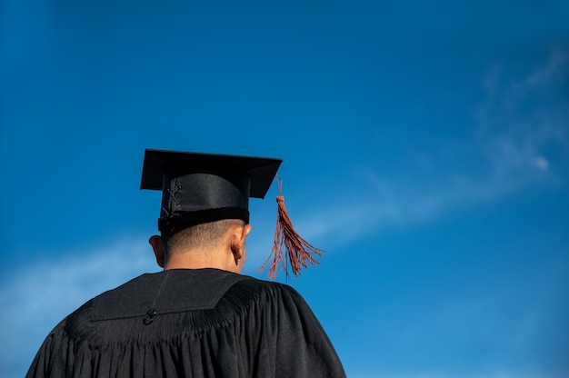 Rear View Of man In Graduation Gown Holding cap Standing Against Sky