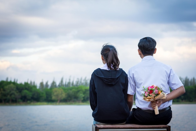 Rear view of man hiding bouquet while sitting with girlfriend on pier against sky