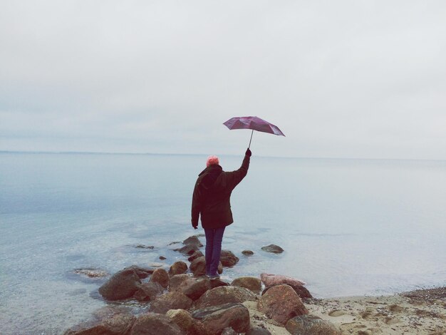 Rear view of man holding aloft umbrella while standing on rocks at beach against sky