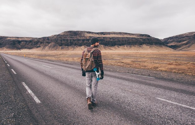 Photo rear view of man holding skateboard walking on road against mountain