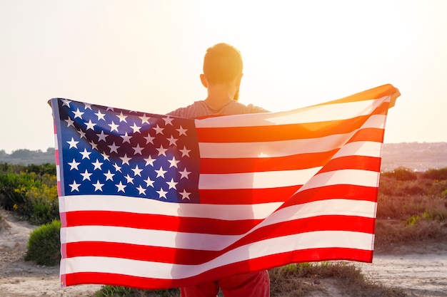 Rear view of man holding waving usa american flag against\
sunset sky outdoor. independence day of united states of america.\
concept of american patriotic people