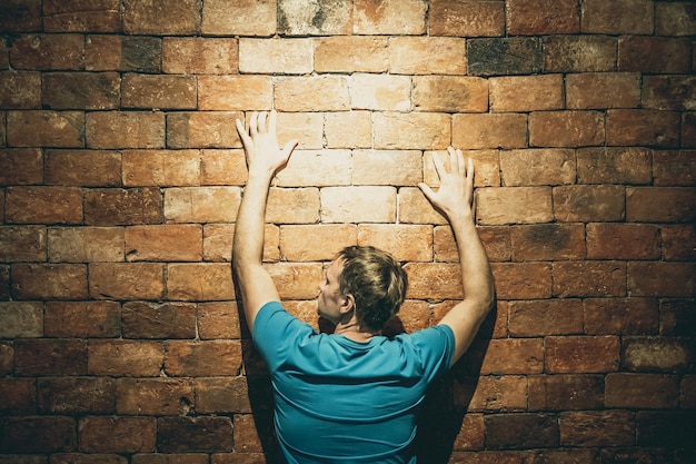 Rear view man under life crisis problems stand facing brown\
brick wall hands raised up on wall wants to climb overcome obstacle\
reach a goal leader motivation looking up left