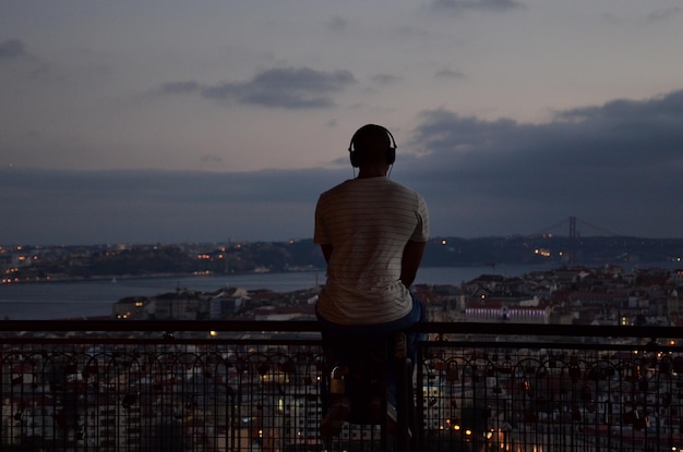 夕暮れの雲の空に照らされた都市風景のレールに座って音楽を聴いている男の後ろの景色
