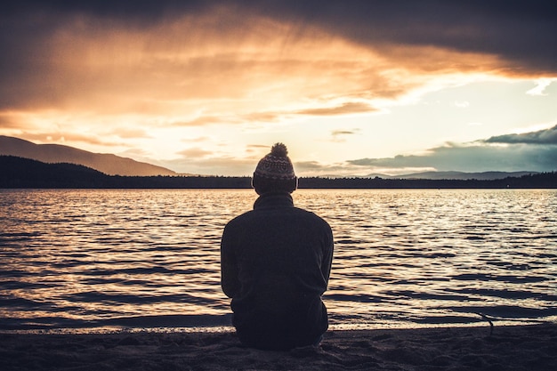 Rear view of man looking at lake against sky during sunset