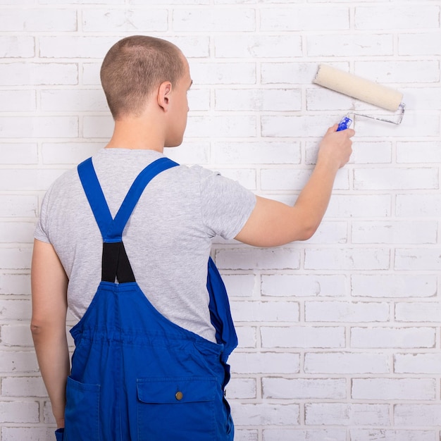 Vista posteriore del pittore uomo in abbigliamento da lavoro dipinto muro di mattoni bianchi con rullo di vernice