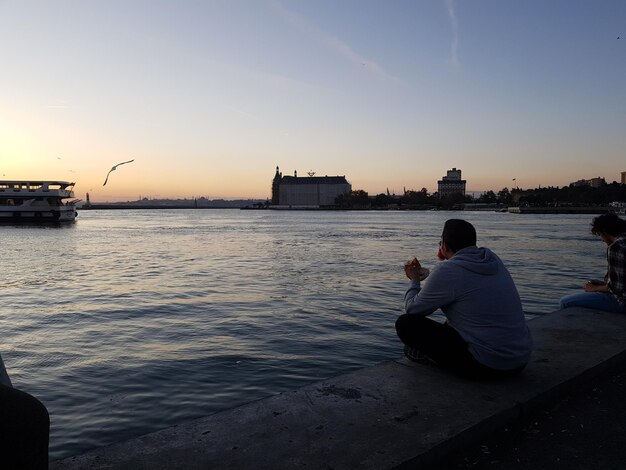 Rear view of man photographing while sitting on river against sky during sunset