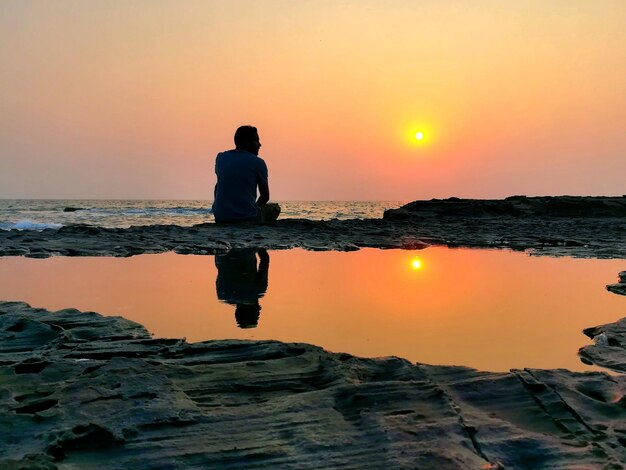 Foto vista posteriore di un uomo seduto sulla spiaggia durante il tramonto con il riflesso in una piccola pozzanghera