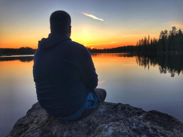 Фотография задний вид человека, сидящего на скале над озером на фоне неба во время захода солнца