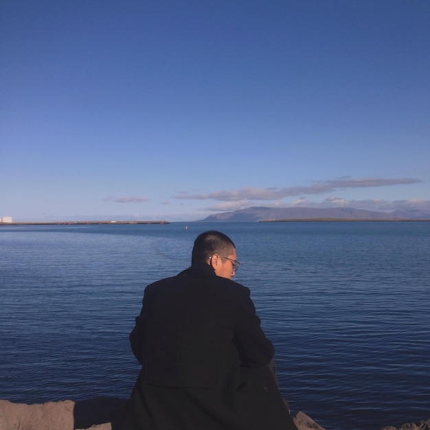 Rear view of man sitting on shore against sky