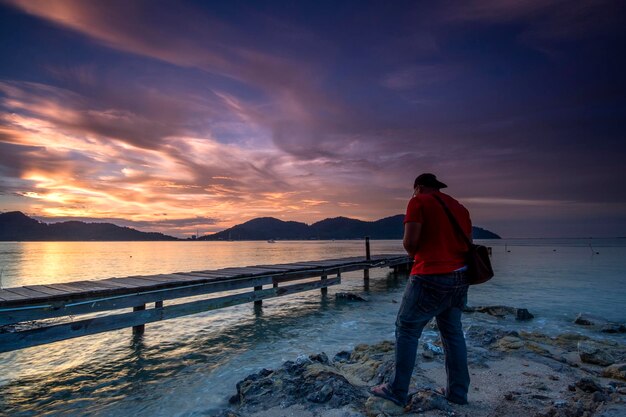 Rear view of man standing at beach against sky during sunset