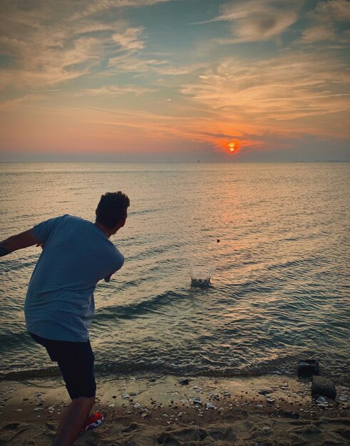 Rear view of man standing at beach against sky during sunset