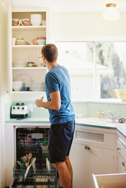 Rear view of a man standing in front of a cupboard with crockery in a kitchen