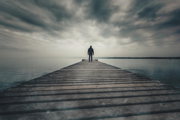 Rear view of man standing on jetty over river against cloudy sky