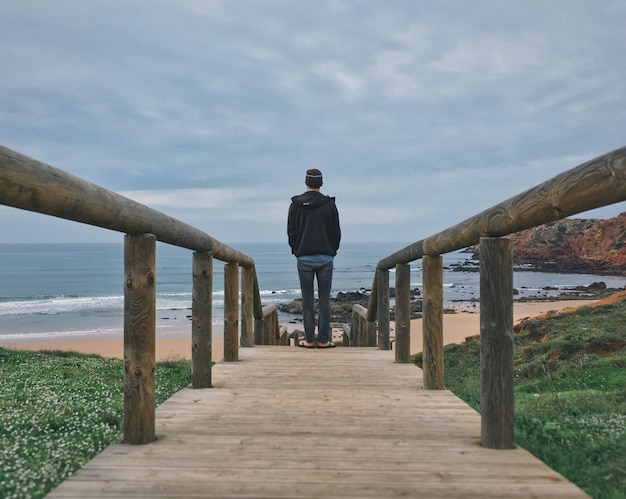 Rear view of man standing on pier at beach against cloudy sky