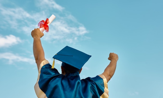 Vista posteriore dell'uomo che lancia le mani in alto un certificato e un berretto in aria, il giorno della laurea sullo sfondo del cielo