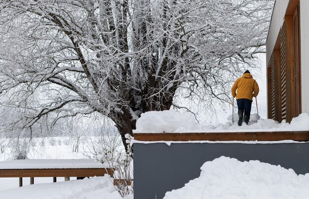 家のテラスで雪を掃除する雪のシャベルを持つ男の背面図