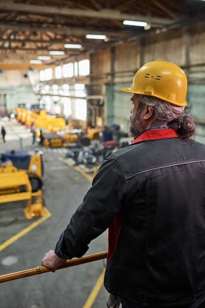 バルコニーに立って工場での生産プロセスを見ている作業用ヘルメットの成熟した職長の背面図