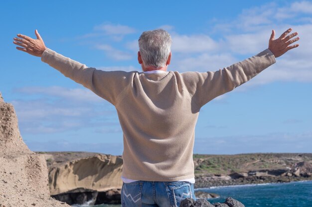 Rear view of mature man enjoying a beautiful day at sea standing on the cliff with outstretched arms