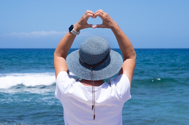 Rear view of mature woman with blue hat looking at horizon over water making heart shape with hands